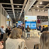 Auf dem Bild ist der Rückansicht von zwei Frauen zu sehen, welche sich einen Vortrag anhören. Vorne hält ein Redner vor seinem Messestand eine Ansprache.