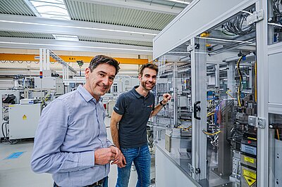 Zwei Männer stehen in einem hellen Industrie- oder Laborraum vor einer komplexen technischen Anlage mit Kabeln und Steuergeräten