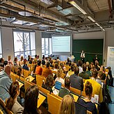 Vortragende steht vor einer Leinwand und spricht zu einem Publikum in einem Hörsaal mit Holzsitzreihen und großen Fenstern