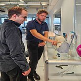 Auf dem Bild sind zwei Herren zusehen. Einer der Herren steht vor einem 3D Modell und scheint die Funktionen zu erklären.