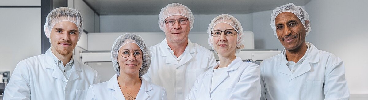 Fünf Personen in weißen Laborkitteln und Handschuhen stehen in einem Laborumfeld