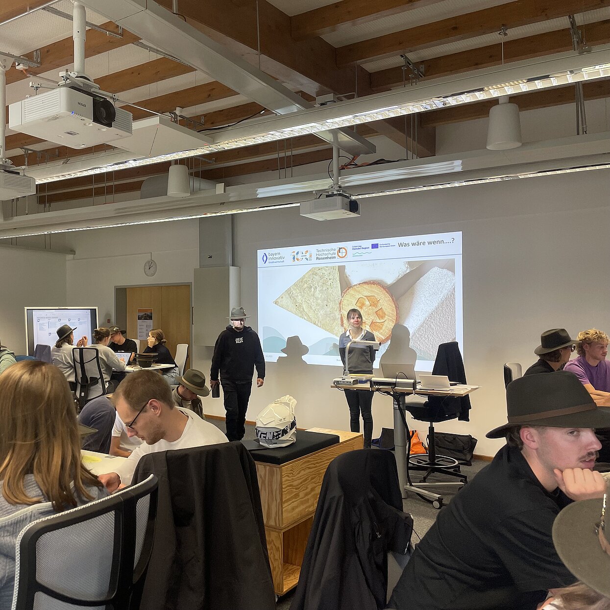 Studierende, einige mit grünen Hüten sitzen in einem Vortragsraum und hören einer Präsentation zu. 