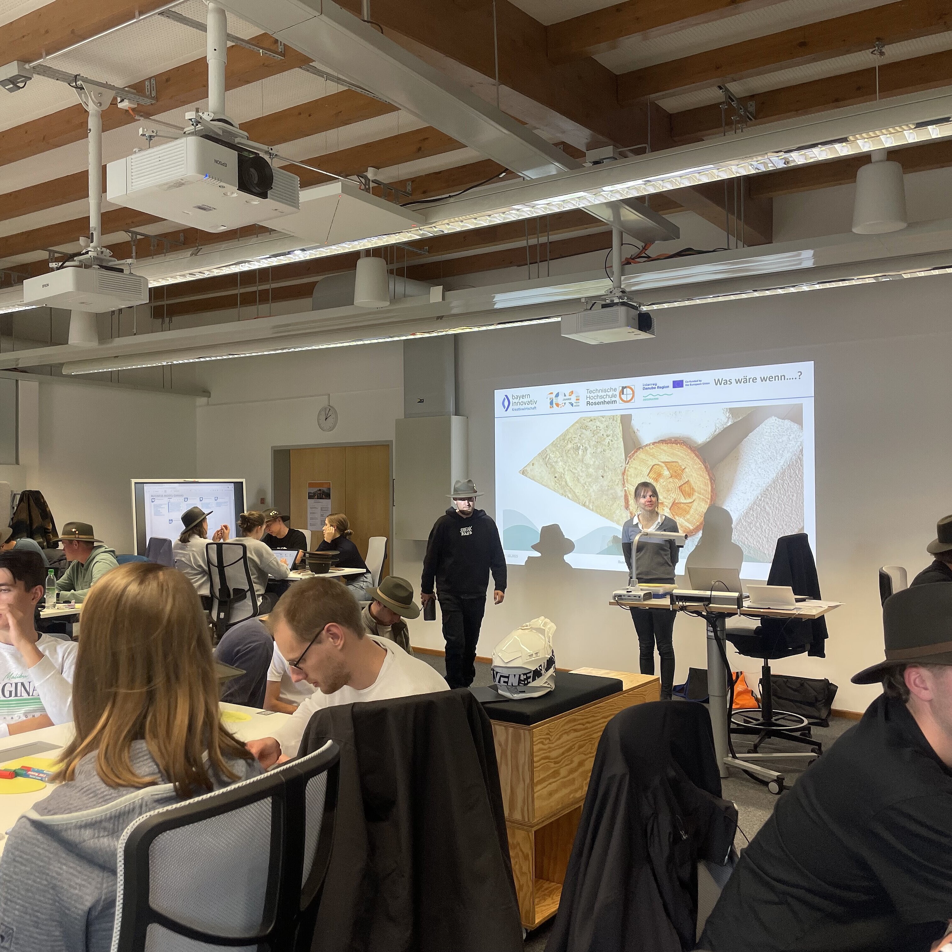 Studierende, einige mit grünen Hüten sitzen in einem Vortragsraum und hören einer Präsentation zu. 