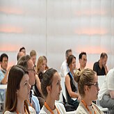 Eine Gruppe von Personen sitzt in einem Raum, der wie ein Klassen- oder Vorlesungssaal wirkt. Alle blicken nach vorn, als würden sie einer Präsentation oder einem Vortrag folgen