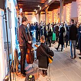 Das Bild zeigt mehrere Personen, die um eine Band herumstehen. Der Raum ist orange beleuchtet, die Personen sind formell gekleidet