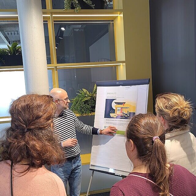 Ein Mann steht vor einem Schreibboard und erzählt etwas. Um ihn herum stehen drei Frauen. 