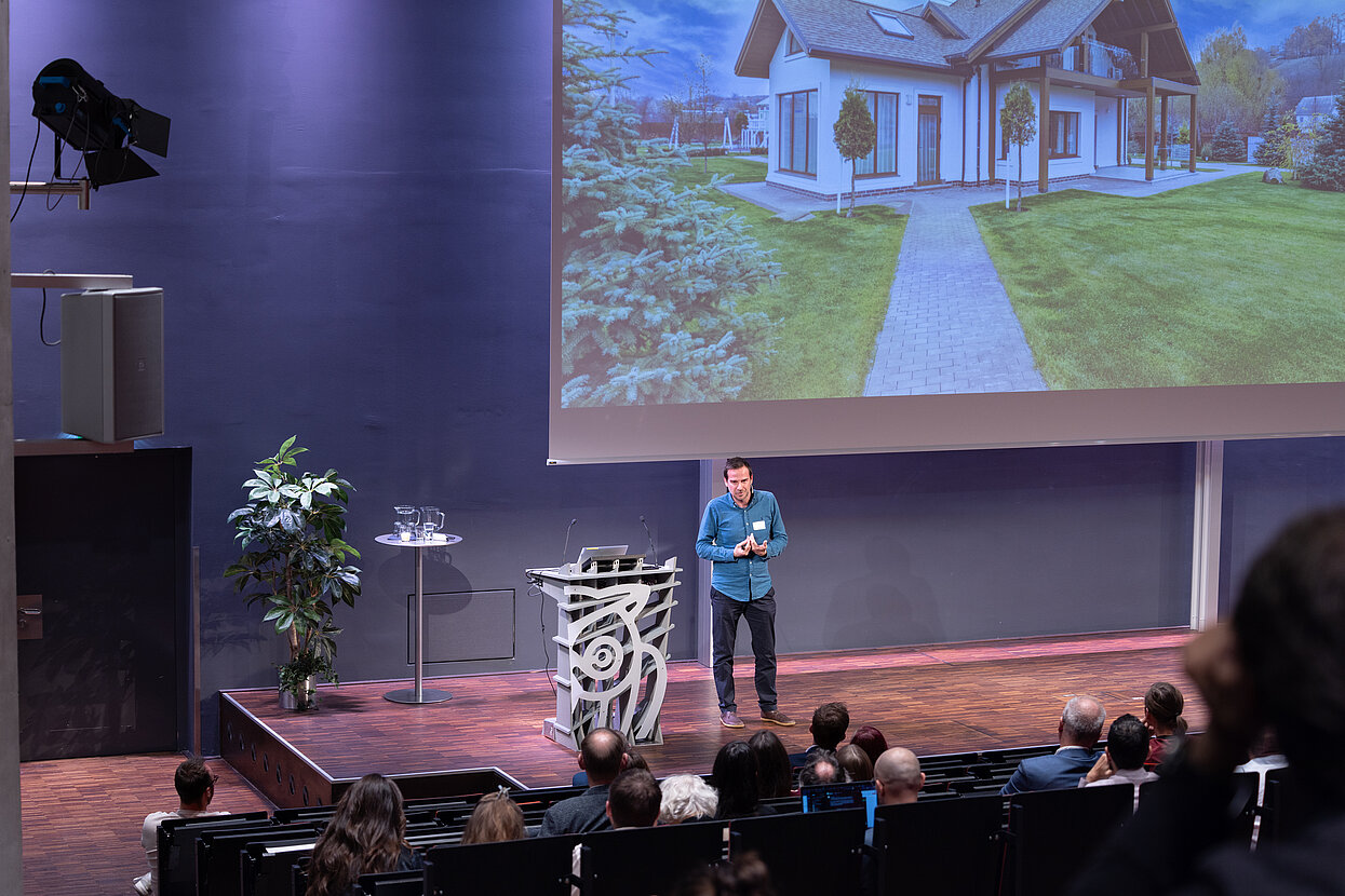 Podium im Hörsaal und Referent auf der Bühne, hinter ihm eine Präsentation auf der Leinwand. 