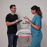 Eine Frau in medizinischer Arbeitskleidung spricht mit einem Mann in schwarzem T-Shirt, der ein Klemmbrett hält, vor einem medizinischen Gerät mit Kabeln an der Wand.