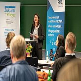 Frau steht vor Publikum und hält Papiere in einem Raum mit Bannern zu Pflege und Innovationen