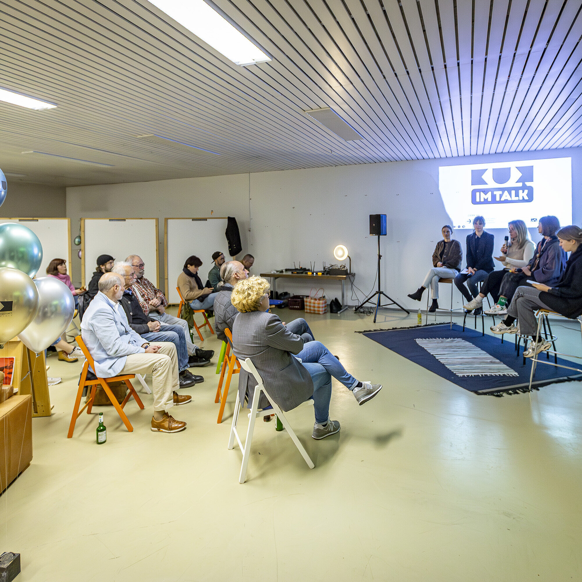 Auf dem Bild sitzt ein fünfköpfiges Panel, darunter Charlotte Stegmayer, Projektmanagerin im Innovationsnetzwerk Kreativwirtschaft der Bayern Innovativ GmbH, auf hohen Hockern vor einem Publikum. Hinter dem Panel ist eine Projektion mit dem Titel ‚KUK im Talk‘ zu sehen. 