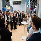 Frau spricht vor einer Gruppe von Menschen in einem hellen, modernen Messestand mit weißen Wänden und Stehtischen