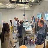 Die Bildaufnahme zeigt eine Gruppe von Personen bei einer geführten Besichtigung in einem Hightech-Labor. Im Mittelpunkt steht ein 3D-Drucksystem für medizinische Anwendungen, das von einem Experten erläutert wird. Die Teilnehmenden hören aufmerksam zu und erhalten Einblicke in den praktischen Einsatz additiver Fertigungstechnologien in der Medizintechnik.