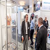 das Bild zeigt eine Gruppe von Personen auf einer Messe oder Ausstellung. Im Vordergrund befinden sich Vitrinen mit ausgestellten Objekten, im Hintergrund sind verschiedene Stände und Banner zu sehen.