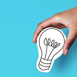 Eine Hand hält eine Kartonfigur einer Glühbirne, während die andere Hand eine Figur eines Geldsacks hält