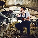 Ein Bauer kniet Lachend im Stall neben einer Kuh und hält ein Tablet in der Hand. 