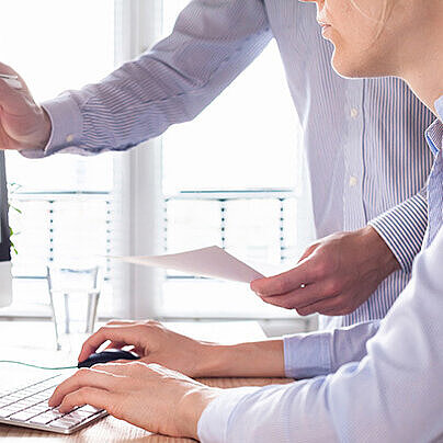 Das Bild zeigt zwei Personen an einem Schreibtisch, die gemeinsam Daten und Diagramme auf einem Computerbildschirm besprechen.