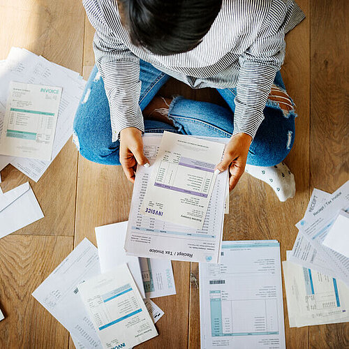 Person sitzt auf dem Boden und sortiert Rechnungen und Belege.