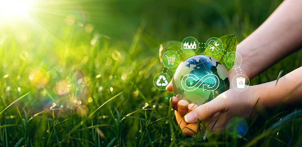 Ein Paar Hände hält sanft einen kleinen Globus in Grün- und Blautönen, der die Erde symbolisiert. Der Hintergrund besteht aus üppigem grünen Gras, durch das von der linken Seite Sonnenlicht strömt und eine helle und lebendige Atmosphäre schafft. Um den Globus herum sind kleine leuchtende Partikel zu sehen, die dem Bild eine magische und friedliche Stimmung verleihen.