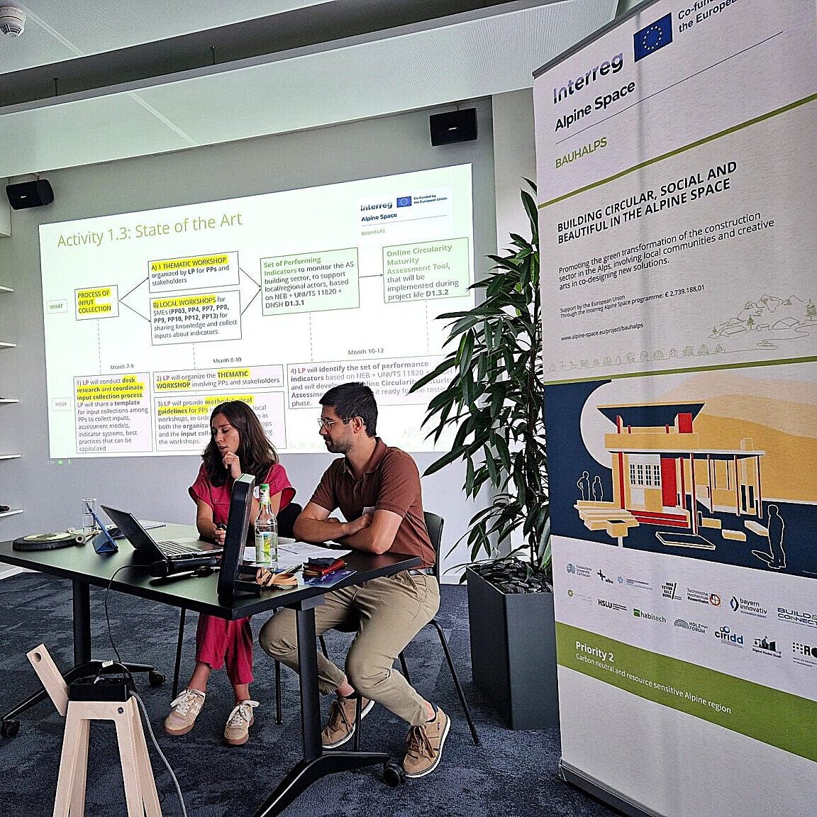 Zwei Personen sitzen an einem Rednerpult vor einer Präsentation. 