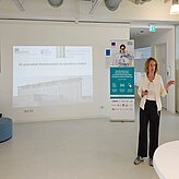 Das Bild zeigt eine Frau, die vor einer Leinwand steht, auf der eine Präsentation über 3D-gedruckten Knochenersatz im operativen Umfeld zu sehen ist. Im Hintergrund ist ein Roll-up-Banner mit Informationen zu einem Projekt sichtbar. Die Frau scheint eine Präsentation zu halten. Der Raum wirkt modern und hell, typisch für ein Büro oder eine Konferenzumgebung. 
