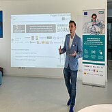 Auf dem Bild ist ein Herr in blauem Sakko und weißem Hemd zu sehen, welcher einen Vortrag zu halten scheint. Im Hintergrund ist auf der Wand eine Präsentation, sowie ein RollUp zu sehen.