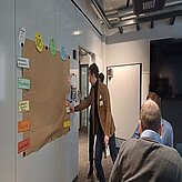 Auf diesem Bild ist eine Gruppe von Menschen in einem Raum mit weißen Wänden und einem Whiteboard zu sehen. Eine Person steht in der Nähe eines großen Stücks braunen Papiers, das an der Wand befestigt ist und von bunten Haftnotizen umgeben ist. 