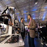 Tobias Gotthardt, Staatssekretär, Bayerisches Staatsministerium für Wirtschaft, Landesentwicklung und Energie und eine weitere Person stehen an einem Messestand und unterhalten sich. Im Vordergrund ist eine technische Anlage zu sehen.