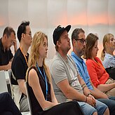 Eine Gruppe von Personen sitzt in Reihen und scheint an einer Veranstaltung oder Präsentation teilzunehmen. Alle blicken nach vorn und wirken aufmerksam