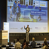 Vortragsbühne auf der ein Referent gerade einen Vortrag hält im Hintergrund ist eine große Leinwand zu sehen mit dem Veranstaltungstitelbild.